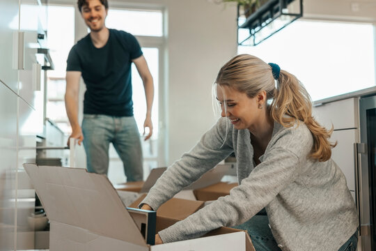 Smiling young woman unpacking things from cardboard box in kitchen at new apartment