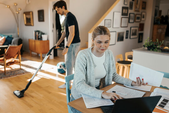 High angle view of young female freelancer doing accountancy while boyfriend vacuuming floor in background at home