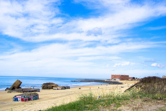 View of sandy beach with weathered bunkers adorned with graffiti under a vast blue sky dotted with fluffy clouds, contrasting with the calm ocean waters, Capbreton, France.