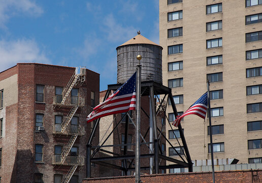 View of American flags fluttering beneath a weathered water tower, amidst brick buildings under a bright sky, a blend of urban charm, New York, New York, United States.