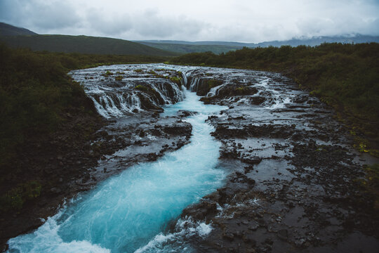 View of glacial blue water cascades Br&uacute;ar&aacute;rfoss over dark, rugged rocks amidst green vegetation under a clouded sky, South Iceland, Iceland.