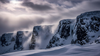 dramatic winter scene with snow covered cliffs and dramatic clouds with sunlight breaking through illuminating mist and windblown snow