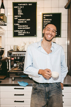 Portrait of smiling male barista holding coffee cup standing near counter in cafe