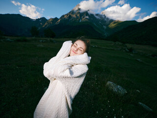 Naklejka premium Softly smiling woman in a thick white sweater hugs herself in a green meadow, distant mountains and a bright blue sky forming a peaceful outdoor scene, conveying warmth, comfort, and connection with
