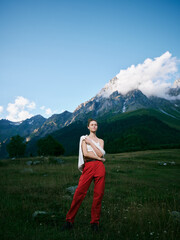 Naklejka premium Mountain landscape, outdoor fashion on a model in a grassy field with red pants and white top, dramatic sky and distant peaks, nature and style in color