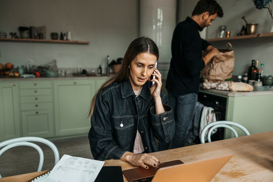 Woman talking on phone while using laptop and doing home finances sitting at table