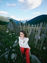 Naklejka premium Woman sits beside a weathered fence in a mountainous countryside landscape, thoughtful expression, casual white sweater and red pants, capturing a serene outdoor moment