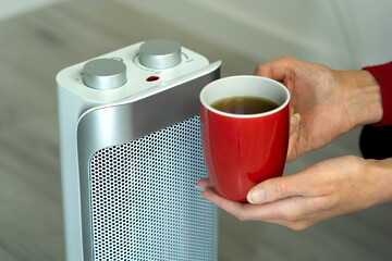 close-up of a cold woman's hands holding a cup of hot tea while warming herself with a portable fan heater (space heater). Energy crisis, icy winter
