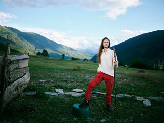 Naklejka premium Woman standing with a walking stick in a wide alpine meadow, mountains in the background, clear sky, fence on the left, rugged terrain, casual outdoor gear, calm countryside scene, exploring nature