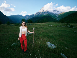 Naklejka premium Woman in red pants holding a wooden staff stands in a lush meadow with distant mountains under a bright sky, capturing an expansive outdoor landscape and a sense of adventure