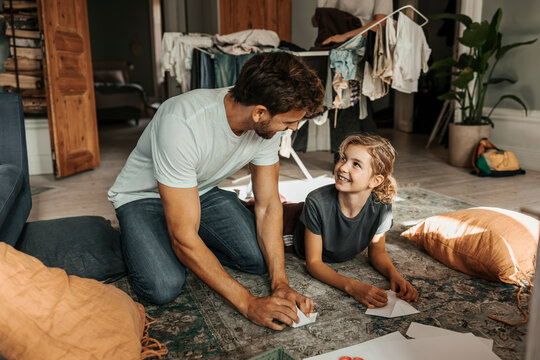 Smiling girl making paper boat with father while lying on carpet in home