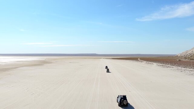 Aerial view of vehicles traverse the vast, flat terrain near Tuzbair Lake, a striking contrast of light sand and blue sky, Mangystau Region, Kazakhstan.