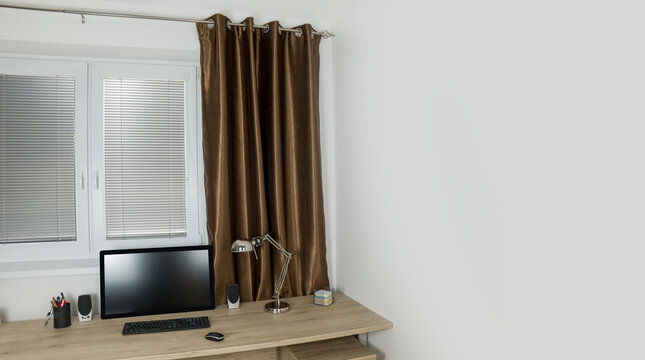 Desk with a monitor keybord and lamp. White plastic window with a blinds. Brown welvet curtain on the rod