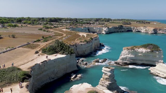 Aerial view of rocky cliffs meeting the turquoise sea, contrasted with earthy land and paths, creating a vibrant coastal scene, Sant'Andrea, Apulia, Italy.