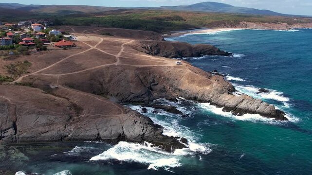 Aerial view of a rugged coastline where turquoise waters crash against dark rocks, meeting dry grassy headlands near a small village, Sinemorets, Bulgaria.