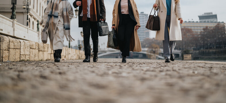 Four business people walk along a city sidewalk in winter coats, carrying bags. A urban street scene shows professionals on a chilly day, dressed for work.