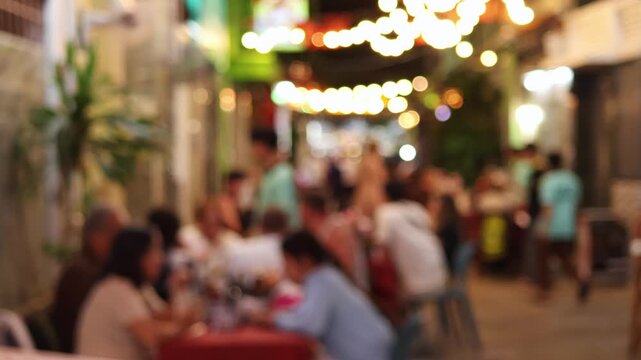 A blurred view of a street restaurant in the evening, decorated with garlands. People are sitting at tables, enjoying food and drinks, and cozy atmosphere. Bokeh real time motion video.