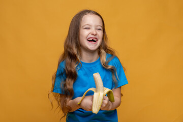 Happy funny laughing little girl without a baby tooth, wearing casual blue t-shirt holds ripe banana in her hands on yellow background, copy space. High quality photo. Children snack concept.