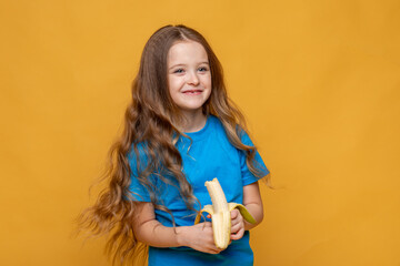 Happy funny laughing little girl without a baby tooth, wearing casual blue t-shirt holds ripe banana in her hands on yellow background, copy space. High quality photo. Children snack concept.