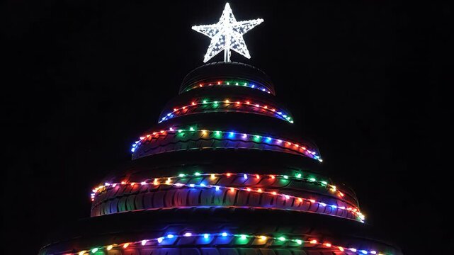 High contrast isolated view of a rotating Christmas tree structure assembled from black rubber tires, crowned with a metallic star and wrapped in continuous glowing lights crowned, rubber, view