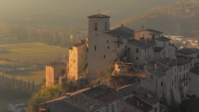 Aerial view of Casteldilago's ancient stone tower and houses nestled on a hilltop, bathed in a soft, golden light, Umbria, Italy.