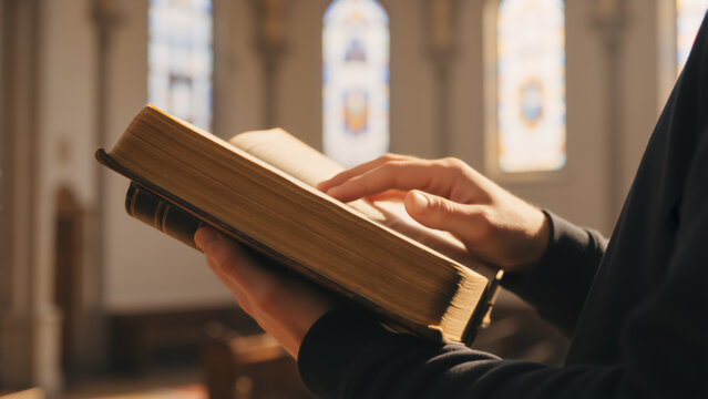 Close-up of hands holding and reading the Holy Bible. Person studying scripture in a sunlit church with stained glass windows