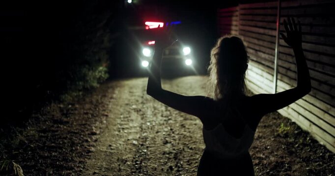 Girl stands with hands raised in front of police silhouette on a dark dirt road at night with flashing lights illuminating the scene