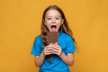 Funny little girl in blue t-shirt holding chocolate bar with nuts on yellow background, closeup. The concept of children's happiness and sweets addiction