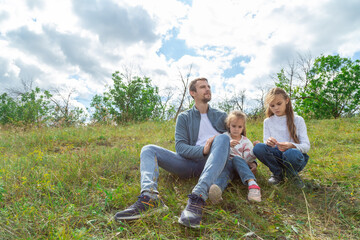 Fototapeta premium Father and daughters sitting on top of hill and enjoy beautiful scenery of green hills under blue cloudy sky.