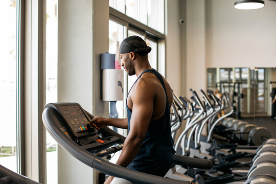 Athletic man adjusting treadmill settings in gym. Muscular black man adjusting treadmill interface in a modern gym. - Powered by Adobe