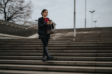 A stylish young woman walks down stairs, looking at her phone in an urban, moody setting. © qunica.com
