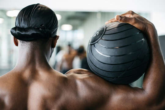 Athletic man holding medicine ball from behind in gym. Rear view of an athletic man holding a medicine ball during functional strength training in a modern gym.