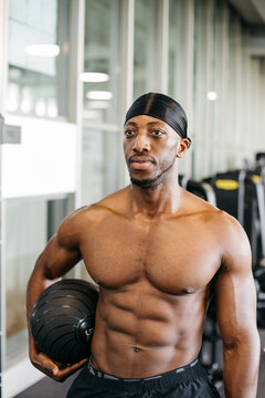 Muscular male athlete holding weighted ball at gym. Shirtless fit man with strong upper body holding a weighted training ball during workout in modern gym.