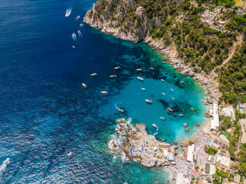 Aerial view of boats dotting the turquoise waters near Marina Piccola, contrasting with the rugged cliffs and lush greenery above, Capri, Campania, Italy.