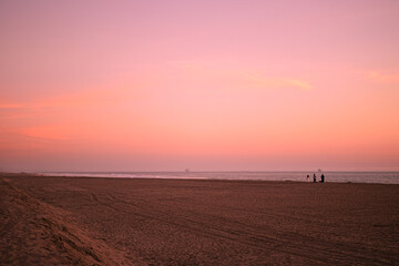 Fototapeta premium Soft pink sunset over a wide sandy beach in Sunset Beach, California, with calm ocean waves and distant silhouettes creating a peaceful coastal scene.