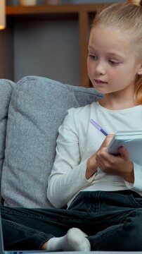 Caucasian kid child girl daughter e-learning class on sofa writing notes raising hand answer to teacher write study and looking laptop during virtual online lesson in cozy living room evening home
