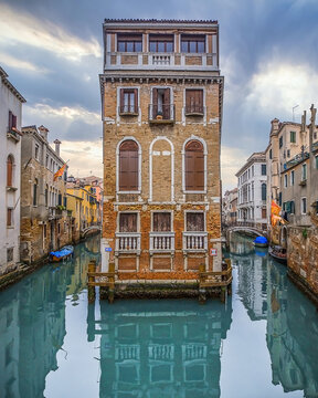 View of a tall, slender, aged building with rusticated stone and arched windows rising from the canal's shimmering turquoise waters, Venice, Veneto, Italy.