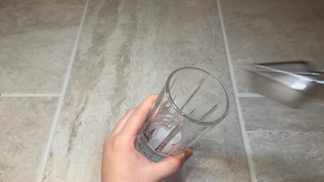 Female hands place ice cubes into a glass using plastic ice tongs. Simple bar preparation action shown from above on a neutral surface, clean and minimal beverage-making concept.