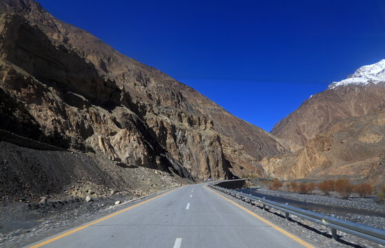 View of a seemingly endless asphalt road winding through towering, craggy mountains under a crisp blue sky, hinting at the rugged beauty of the landscape, Passu, Gilgit Baltistan, Pakistan.