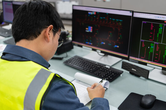 Electrical engineer verifying recorded operational data by crosschecking written logs with control screens inside power facility, ensuring accuracy, validation, and reliable electricity distribution.