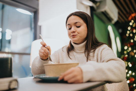 A young woman in a cream sweater eats from a bowl at a cafe table. A festive scene with Christmas tree lights adds warmth and atmosphere.