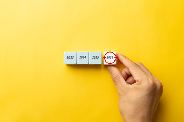 Hand placing 2026 wooden cube with bullseye icon next to year 2023, 2024, and 2025 on yellow background. Concept for business timeline, reaching future targets, and strategic planning for success.