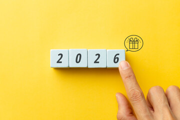 Finger pointing to 2026 wooden cubes with a gift box icon on yellow background. Concept for upcoming year rewards, business bonuses, special offers, and celebrating future success and milestones.