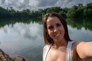A woman is smiling and taking a selfie in front of a lake. The water is calm and the sky is partly cloudy