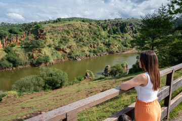 A woman is standing on a wooden fence overlooking a river. The scene is peaceful and serene, with the woman taking in the beautiful view of the water and the surrounding landscape