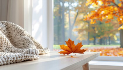 Cozy autumn morning view from a window with a warm knitted blanket and a vibrant orange maple leaf on a wooden surface, overlooking a beautiful fall landscape.