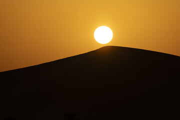 View of the sun blazing over the dark silhouette of a stark desert dune landscape, creating a mesmerizing contrast of light and shadow, Bateen Liwa, Abu Dhabi, United Arab Emirates.