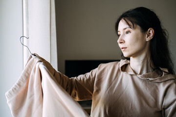 Woman looks at garment on hanger near window. High-quality basics, slow fashion movement, textile sustainability.