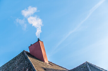 Gekippter rauchender Schornstein mit roter Metallverkleidung an teilweise frostbedecktem Walmdach vor blauem Himmel mit Schleierwolken in Neckartenzlingen.