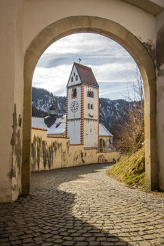 View of aged stone archway framing a clock tower with a red roof against a backdrop of snow-dusted mountains, creating a timeless scene, Fussen, Bavaria, Germany.